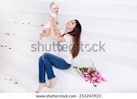 Beautiful and happy mom brunette in white t-shirt and jeans throws up a little smiling baby daughter on a white background with tulips