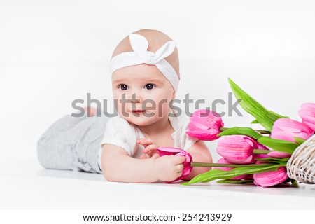 Small very cute wide-eyed smiling baby girl lying on her tummy on a white background in the Studio and keeps handles tulips