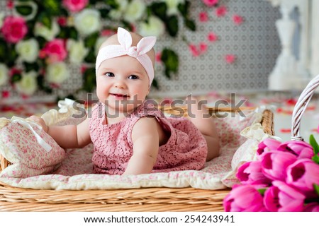 Small very cute wide-eyed smiling baby girl in a pink dress lying in a wicker basket about pink tulips