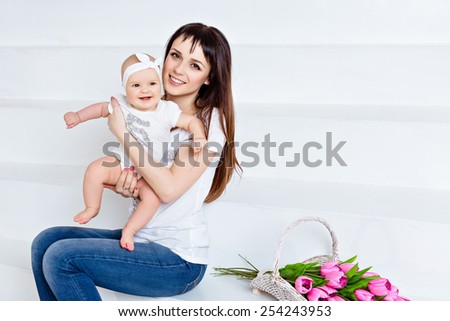 Beautiful and happy mom brunette in white t-shirt and jeans holding a little smiling baby daughter on a white background in the Studio, next to the tulips