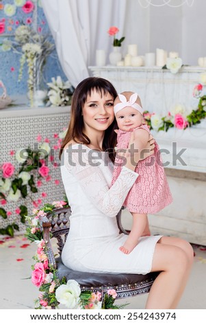 Beautiful and happy mom brunette in a white dress holding on the handles a little girl daughter on the background of the spring interior with flowers