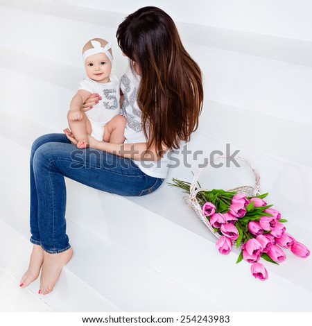 Beautiful and happy mom brunette in white t-shirt and jeans holding on her knees a little smiling baby daughter on a white background with tulips