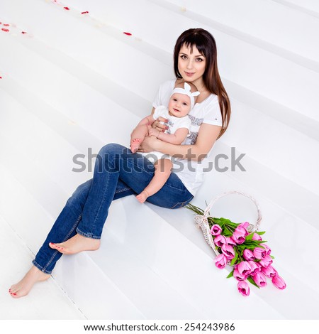 Beautiful and happy mom brunette in white t-shirt and jeans holding on the handles a little smiling baby daughter on a white background with tulips