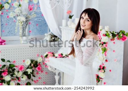 very beautiful sensual brunette girl with long straight hair and is smiling in a white lace dress on the background of spring flower decorations and a white piano