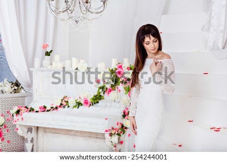 very beautiful sensual brunette girl with long straight hair is in a white lace dress on the background of spring flower decorations and a white piano