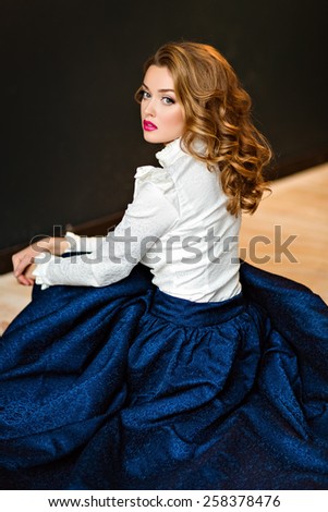 Portrait of a very beautiful sensual glamorous red-haired girl in a white blouse and blue skirt, sitting on the floor in the Studio on a dark background