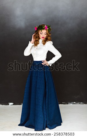 Portrait of a very beautiful sensual glamorous yellow-haired girl in a white blouse and a blue skirt with a wreath of flowers on his head, in the Studio on a dark background, full length