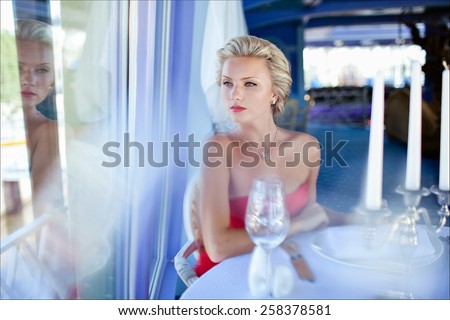 Beautiful elegant exquisite blonde girl in red dress sitting at a table in the restaurant and looks through the glass, reflecting in it