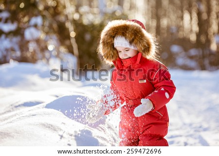 Small very cute girl in a red suit with fur hood plays with snow in winter on the background of the sunset forest
