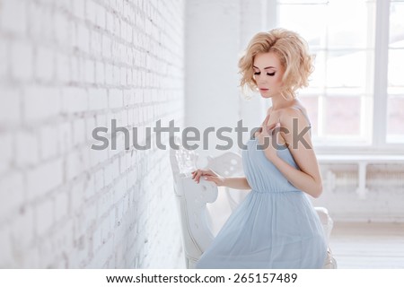 Portrait of a beautiful sensual and sexy blonde girl in a blue dress sitting on a chair against a white brick wall in the Studio