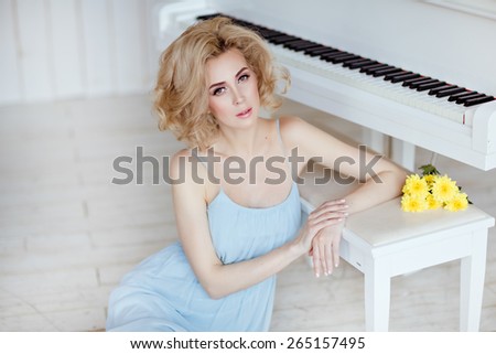 Beautiful sensual and sexy blonde girl in a blue dress sitting at the piano against a white brick wall