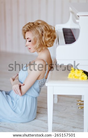 Beautiful sensual and sexy blonde girl in a blue dress sitting at the piano against a white brick wall in the Studio, portrait in profile