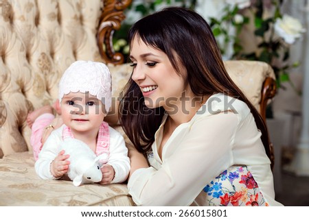 Happy and beautiful mom brunette looks at the charming little daughter baby, in the interiors of the house in beige