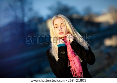 Portrait of beautiful sensual girl with thick blond hair, eyes closed in a black coat and pink scarf fall on a blue background
