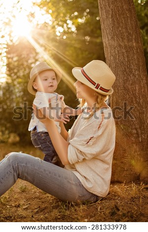 Slender beautiful blonde mom in jeans, a beige shirt and a hat holding a baby boy on the background of the Park at sunset and smiles