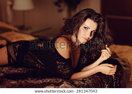 Sexy very beautiful brunette girl with curly hair in black fishnet dress lies on the bed in the interiors of the house