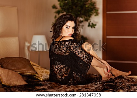 Sexy very beautiful brunette girl with curly hair in black fishnet dress sitting on the bed in the interiors of the house