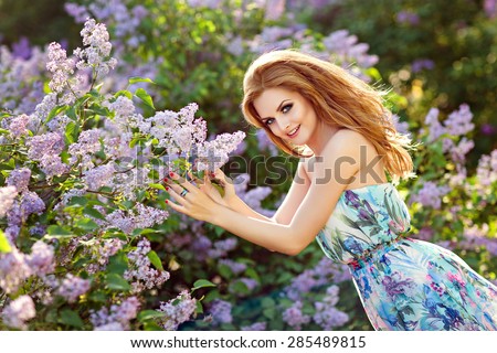 Beautiful glamorous red-haired girl in a bright dress smiling and standing next to a large Bush of lilacs in the summer