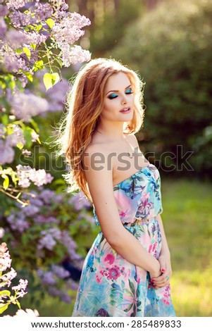 Beautiful glamour girl with long wavy hair and beautiful make-up , in a bright dress stands near a Bush of lilacs in the summer with my eyes closed