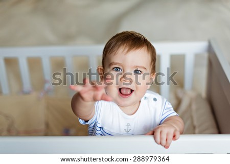 Little cute adorable little blond boy in a striped bodykit is in the nursery with white crib and holds the handle