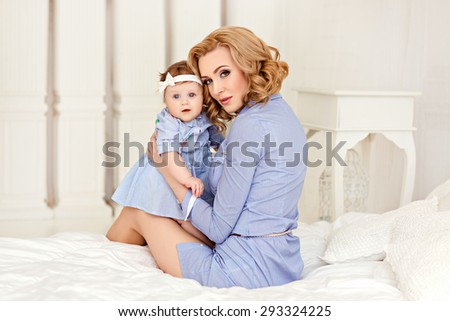 Portrait of a beautiful mom blonde and a little girl baby with blue eyes in blue striped dress sitting on a white bed.