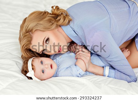 Portrait of a beautiful mom blonde and a little girl baby with blue eyes in a striped blue dress lying on white bed.