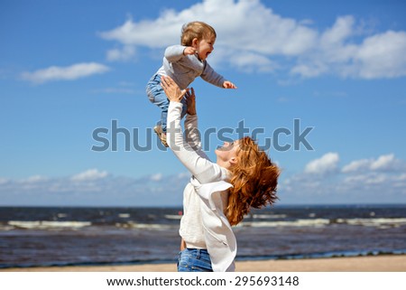 Redheaded mom in jeans and a white sweater throws up his young son against the sea and clouds