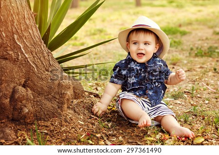 Little adorable baby boy in a straw hat and blue shirt sitting smiling near a tree on earth at sunset in summer