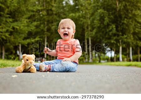 Little boy the kid in blue jeans crying bitterly, sitting on the ground next to a soft toy in the summer