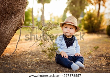 Little adorable baby boy in a straw hat and blue pants sitting with pursed lips, near a tree on earth at sunset in summer