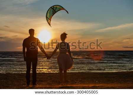 Silhouettes of men and women in the lush short skirt, reaching into the distance on the sand against the sea, the gold of sunset and a flying kite