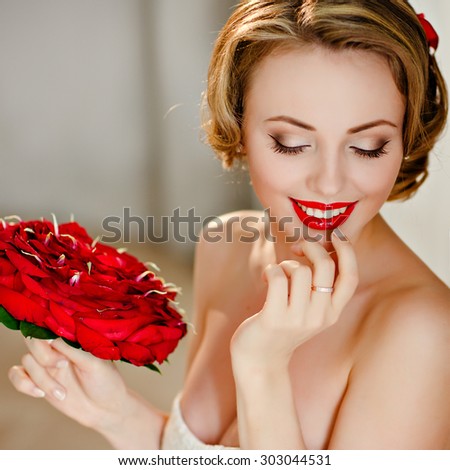 Portrait of a charming girl blonde with beautiful smile and red lipstick, with her eyes closed ,with a bouquet of red roses on the background of the interior, close-up