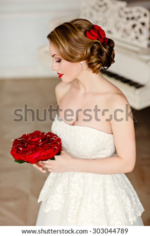 Charming blonde girl with beautiful smile in a white lace dress and a bouquet of red roses is on the background of the interiors in the Studio and looking to the side