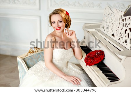 Charming blonde girl with beautiful smile in a white lace dress and a bouquet of red roses sits in an old chair next to the piano on the background of the interior