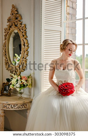 Charming beautiful young blonde girl with red lipstick on my lips, with red roses and white lace dress sits on the window in the interiors of the house, eyes closed