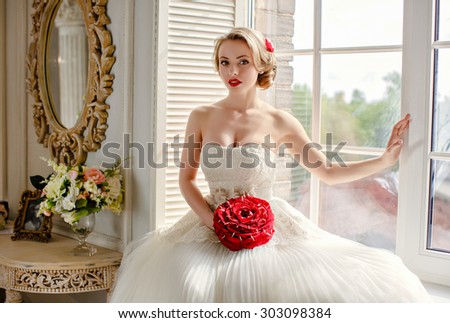 Charming beautiful young blonde girl with red lipstick on my lips, with red roses and white lace dress sits on the window in the interiors of the house