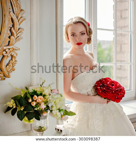Charming beautiful young blonde girl with red lipstick on my lips, with red roses and white lace dress standing at the window in the interiors of the house