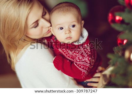 Mom blonde in white sweater kisses little boy the kid in the red sweater at the Christmas tree and garlands in the house