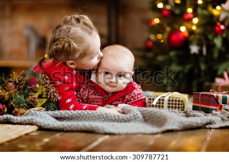 Little boy toddler in a red sweater kisses his older sister, lying together on a cozy blanket on the Christmas tree and garlands in the house next to the gifts