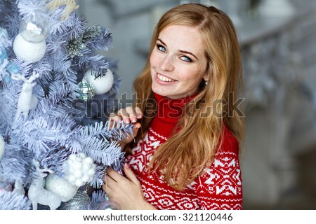 Beautiful red-haired girl with freckles and with blue eyes in a red sweater with white ornaments stands near a tree in the New year and smiles