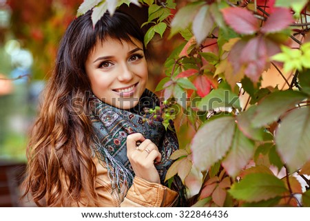 Beautiful and very charming young brunette girl with big brown eyes smiles on the background of red vine leaves in autumn, close-up