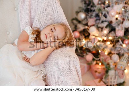 Very cute little girl blonde in white dress sitting on a chair against a background of Christmas trees in the interior of the house