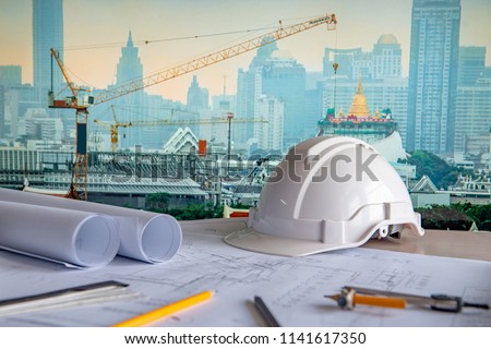 Architectural drawing plan with blueprint rolls, safety helmet and drawing tools on working table. Blurred crane and construction site in Bangkok in the background. Building construction concept