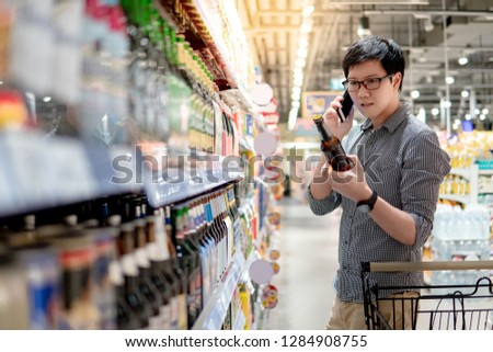 Asian man using phone shopping beer in supermarket. Male shopper with shooping cart choosing beer bottle in grocery store.