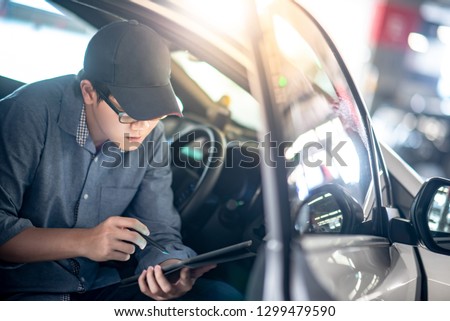 Asian auto mechanic sitting on driver seat checking the car using digital tablet in auto service garage. Mechanical maintenance engineer working in automotive industry. Automobile servicing and repair