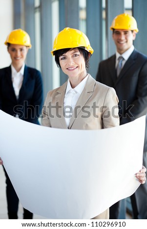 middle aged female architect holding blue print in front of colleagues