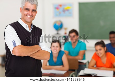 smiling middle aged high school teacher with arms folded standing in front of the class