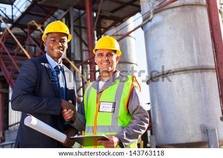 cheerful african refinery manager handshaking with senior worker in front of fuel-tanks