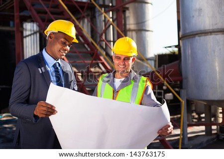 industrial engineers standing in front of a large oil refinery machinery with blueprint on hand