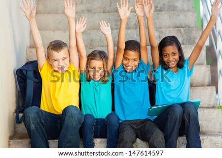 group of happy primary students with hands raised sitting outdoors
